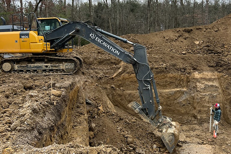 a large excavator digging a hole with a person at the bottom of the hole monitoring the dig